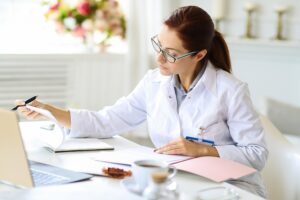 Person wearing a white medical practitioner coat sitting at a desk with an open laptop, holding a pen and reviewing documents. A cup of coffee and a pink folder are on the desk, with a bright background featuring flowers and decorative items.
