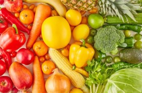 A bright assortment of fresh, whole vegetables arranged closely together, including peppers, carrots, onions, broccoli, corn on the cob, tomatoes, and green leafy vegetables.